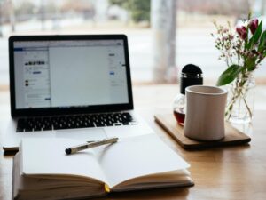 A picture of a laptop computer on a desk with a notebook that is open with a cup of tea to the right of the notebook.